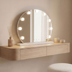 Round white LED vanity mirror with nine bulbs standing on a dressing console beside a reed diffuser