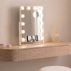 White tilting LED vanity mirror with 12 bulbs and a phone holder on a dressing table, a make-up tutorial playing on the phone